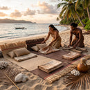 Lifestyle scene of woven lauhala island mats in beige and dark chocolate laid out on a sandy beach under palm trees