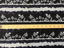 A close-up view of the Lagoon Nights Rayon fabric, displaying a repeating tropical pattern of white palm trees, tiki huts, and sailboats on a dark navy blue background. A wooden ruler is placed horizontally across the center of the fabric to indicate scale, showing that the repeating horizontal bands of the island scenery are approximately 3 to 4 inches in height.