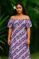 "A woman wearing an off-the-shoulder maxi dress made from the purple and navy Tiare Monstera fabric, posing against a lush green tropical leaf background. She has a white Tiare flower tucked behind her ear, complementing the floral motifs in the textile's traditional Polynesian design."